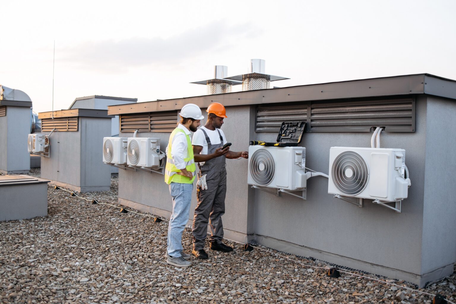 Group of multicultural construction workers on roof inspecting cooling system together using testing tool. Large and gray air conditioning unit with multiple ventilation openings and pipes.